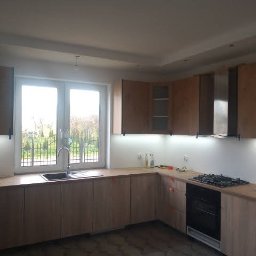 USŁUGI OGÓLNOBUDOWLANE Łukasz Lieske - Jasna kuchnia z drewnianymi szafkami i dużym oknem z widokiem na zieleń, oświetlona listwami LED pod szafkami wiszącymi, z czarną płytą gazową i piekarnikiem.