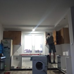 USŁUGI OGÓLNOBUDOWLANE Łukasz Lieske - Wnętrze mieszkania w trakcie montażu szafek kuchennych; widoczny mężczyzna na krześle instalujący górne szafki, pralka stojąca na środku pomieszczenia, okno z widokiem na budynki i drzewa.