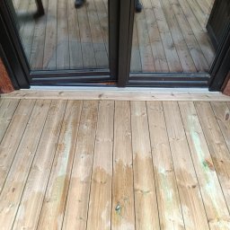 USŁUGI OGÓLNOBUDOWLANE Łukasz Lieske - Jasne deski tarasowe z widocznymi słojami, prowadzące do czarnego, przeszklonego wyjścia. Elementy konstrukcyjne z drewna w kolorze brązowym i zielonym.