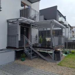 Nowoczesny taras z zadaszeniem i metalowymi schodami, szara kolorystyka, widok z poziomu gruntu. Elementy konstrukcyjne z metalu.