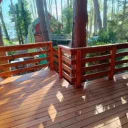 Wykończony taras drewniany z balustradą, otoczony zielenią drzew. Widoczne deski podłogowe i elementy konstrukcyjne, całość w ciepłych, naturalnych odcieniach drewna.