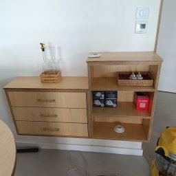 USŁUGI OGÓLNOBUDOWLANE Łukasz Lieske - Drewniana szafka na ścianie z trzema szufladami i otwartymi półkami, na których leżą dekoracje i akcesoria. W tle biała ściana i odkurzacz.