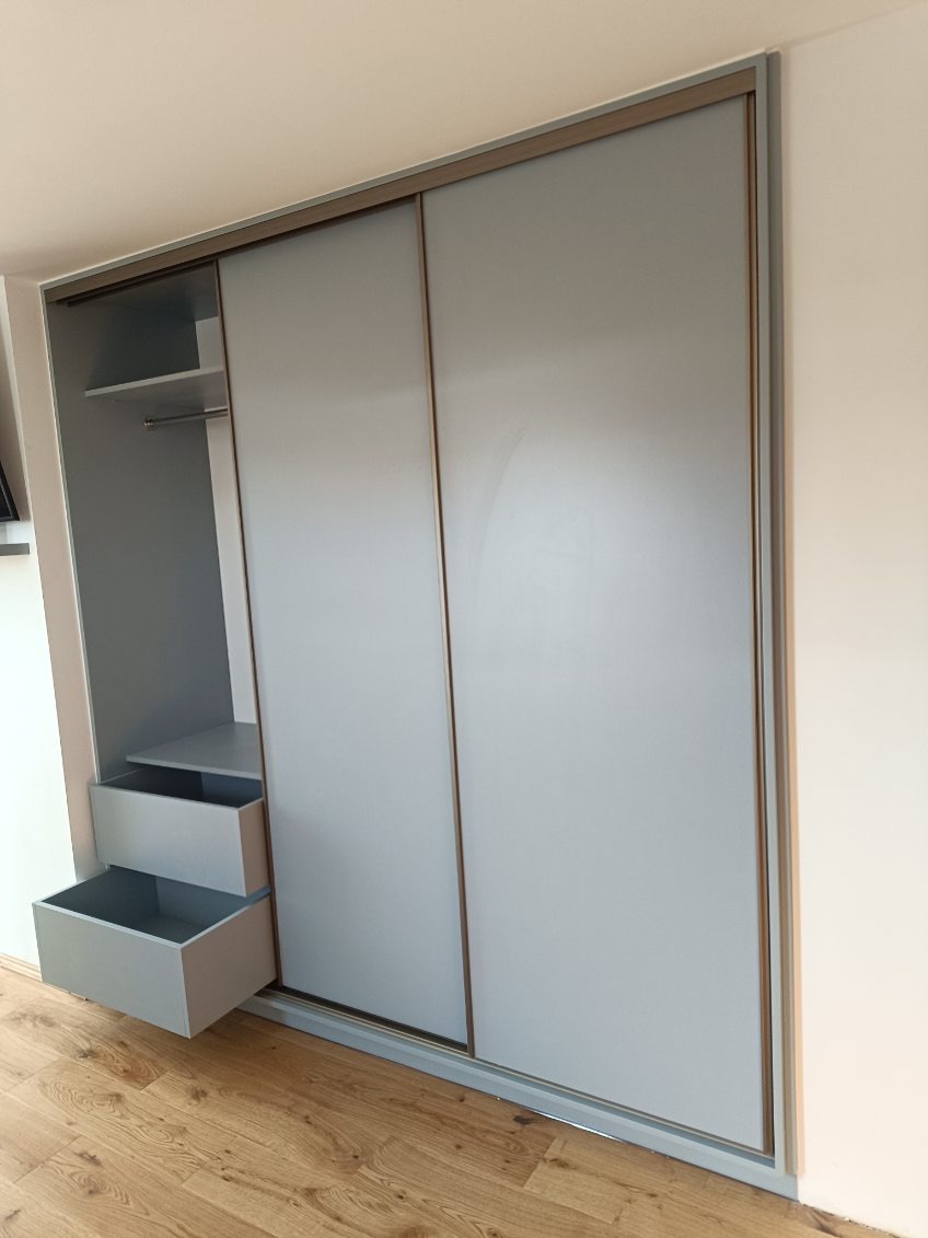 Jasnoszara szafa przesuwna z otwartą częścią z półkami i szufladami, widoczna na tle drewnianej podłogi. Minimalistyczny design, neutralne kolory.