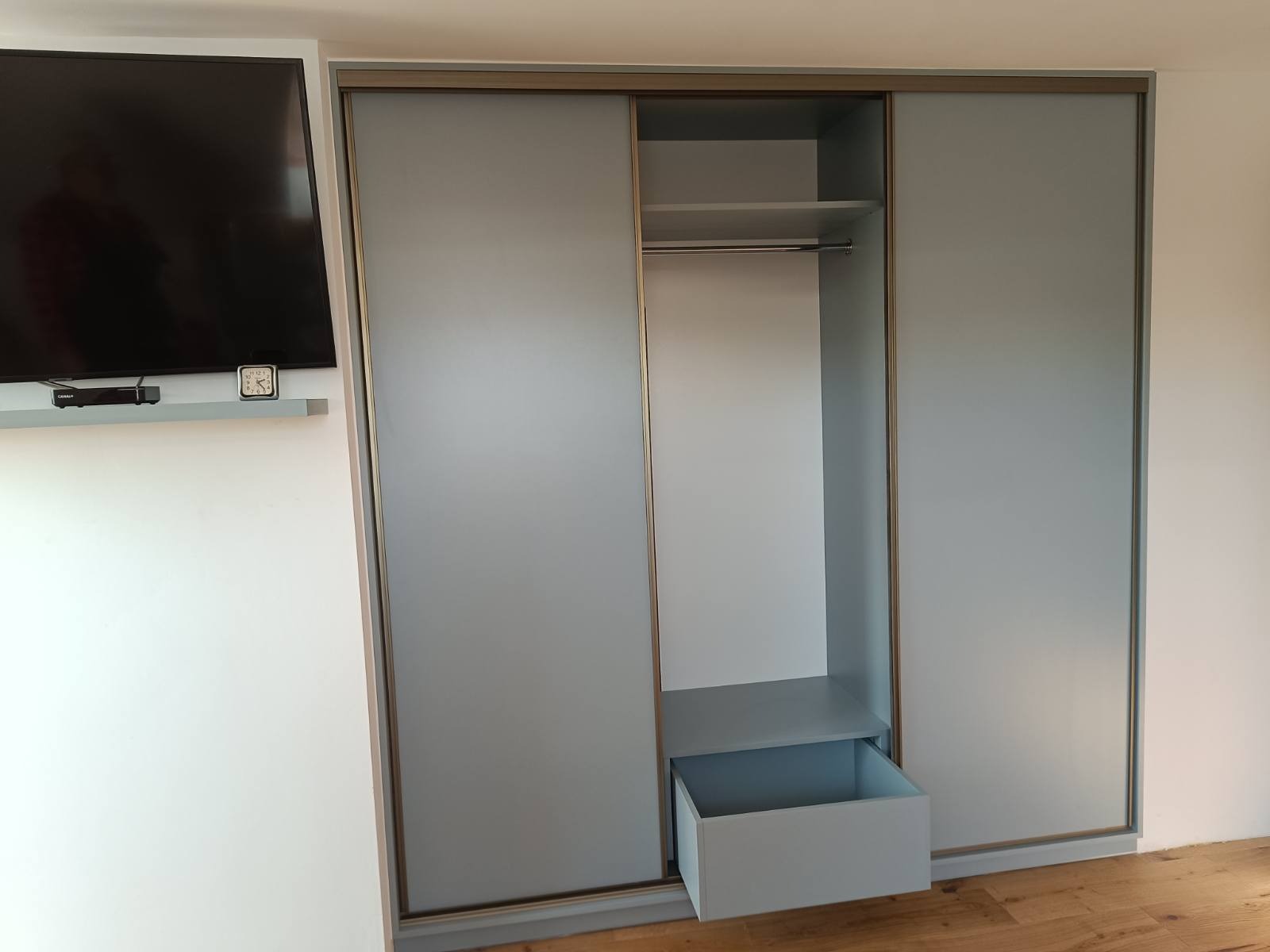 Jasnoszara szafa przesuwna z otwartym frontem i wysuniętą szufladą, widoczna półka i drążek na ubrania. Minimalistyczny design, drewniana podłoga.