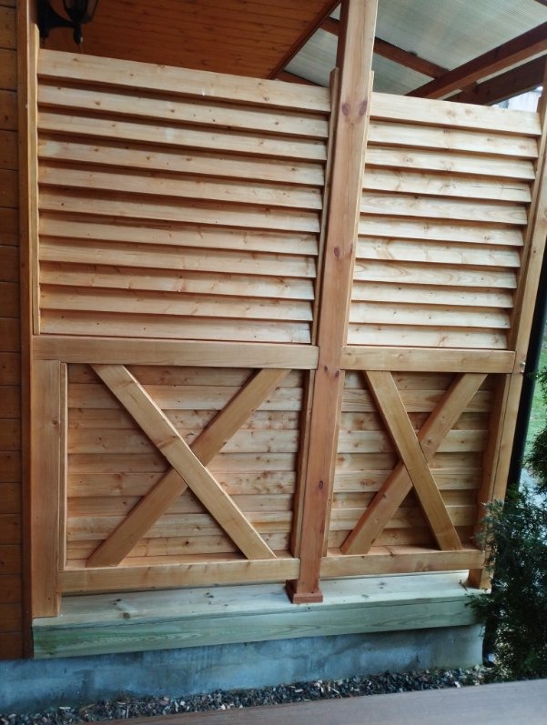 Drewniana konstrukcja z poziomych desek i krzyżowych wzmocnień, prawdopodobnie element pergoli lub altany, z widocznym oświetleniem i fragmentem dachu.