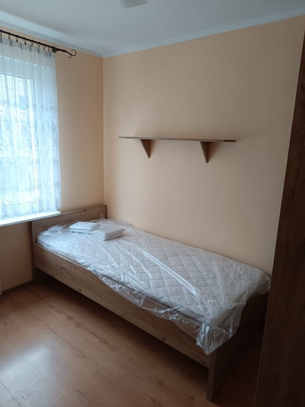 Jasne wnętrze pokoju z pojedynczym łóżkiem z drewnianą ramą, przykrytym folią ochronną, z widocznymi dwiema poduszkami i półką na ścianie.
