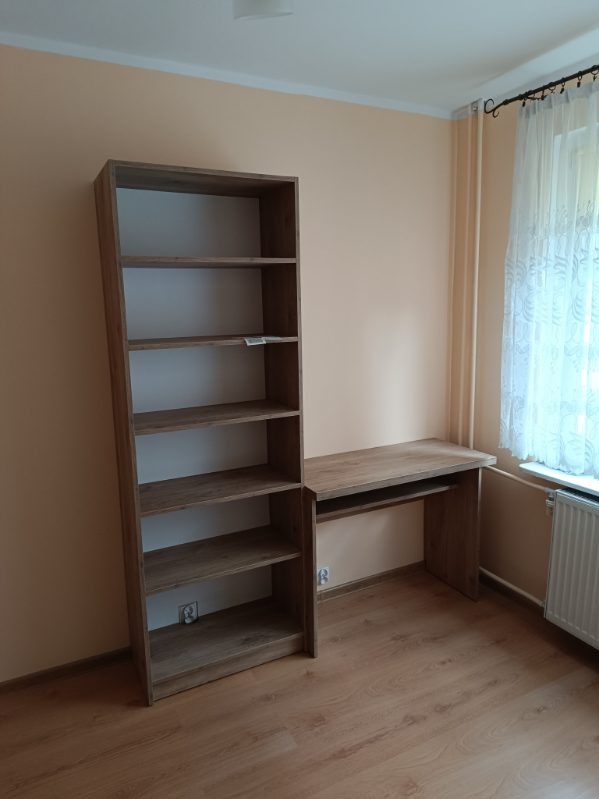 Brązowy regał z półkami i biurko z płyty w pokoju o jasnych ścianach i panelach. Widoczne okno z firanką i grzejnik. Prosta aranżacja wnętrza.