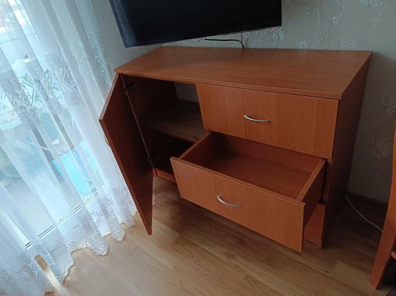 Komoda z drewna w odcieniach brązu, z otwartymi drzwiczkami i szufladą, na tle okna z firanką, widoczna pod telewizorem. Ukazana na tle podłogi z paneli.