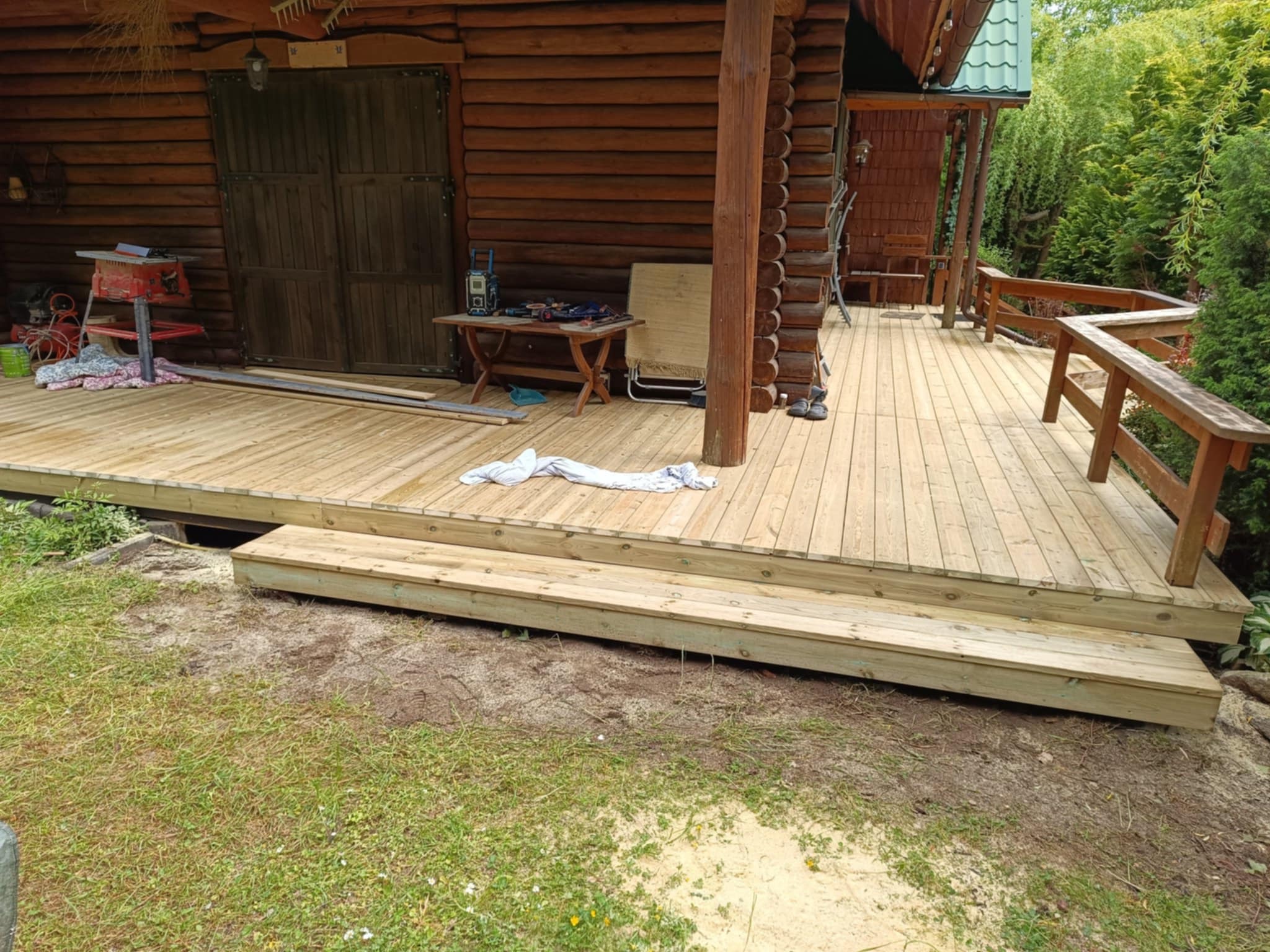 Nowy, drewniany taras z balustradą przy rustykalnym domu z bali, widok z poziomu trawnika. Elementy robocze na tarasie. Jasne drewno, zielone otoczenie.