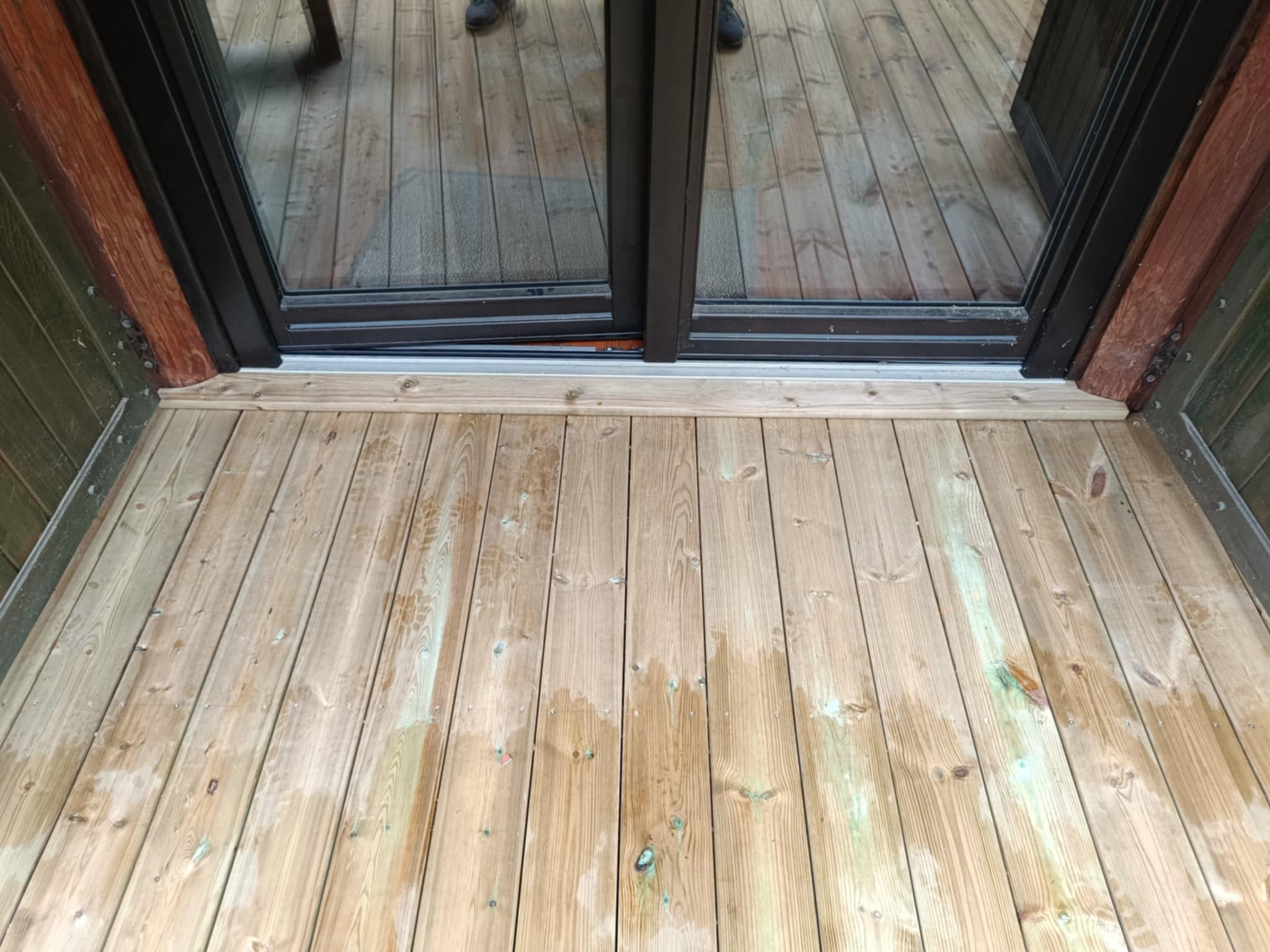 Jasne deski tarasowe z widocznymi słojami, prowadzące do czarnego, przeszklonego wyjścia. Elementy konstrukcyjne z drewna w kolorze brązowym i zielonym.