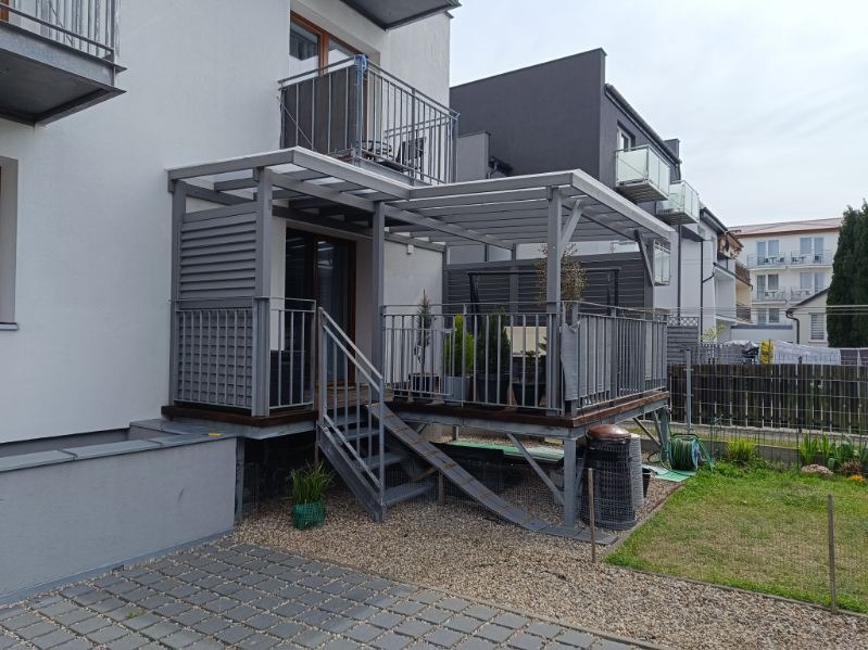 Nowoczesny taras z zadaszeniem i metalowymi schodami, szara kolorystyka, widok z poziomu gruntu. Elementy konstrukcyjne z metalu.