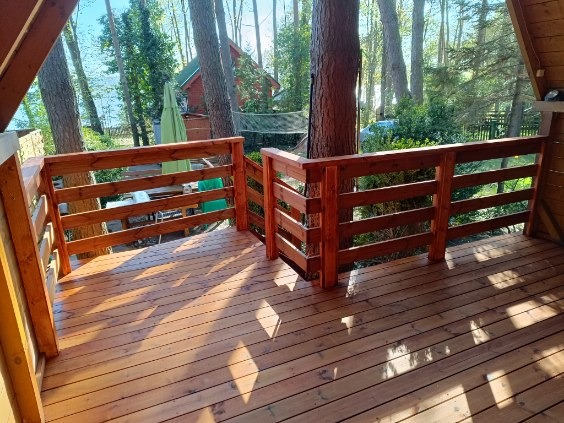 Wykończony taras drewniany z balustradą, otoczony zielenią drzew. Widoczne deski podłogowe i elementy konstrukcyjne, całość w ciepłych, naturalnych odcieniach drewna.