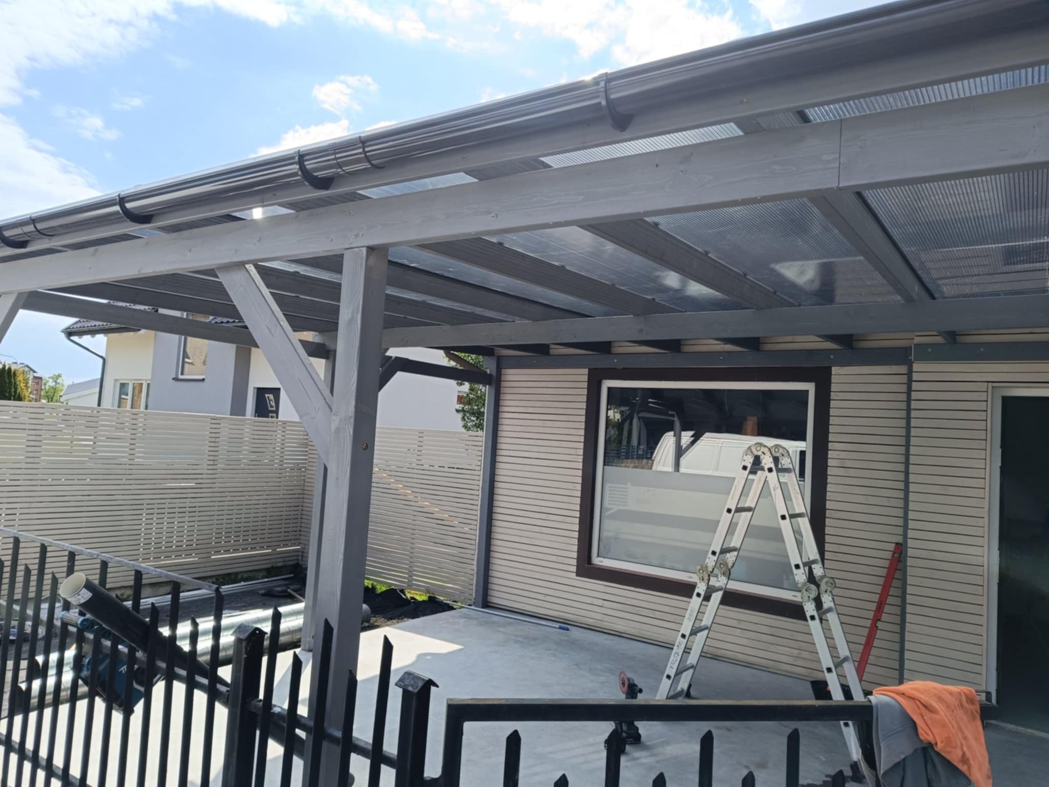 Szara drewniana konstrukcja z zadaszeniem z poliwęglanu, widok z dołu, drabina oparta o ścianę, barierka w tle, jasne niebo.