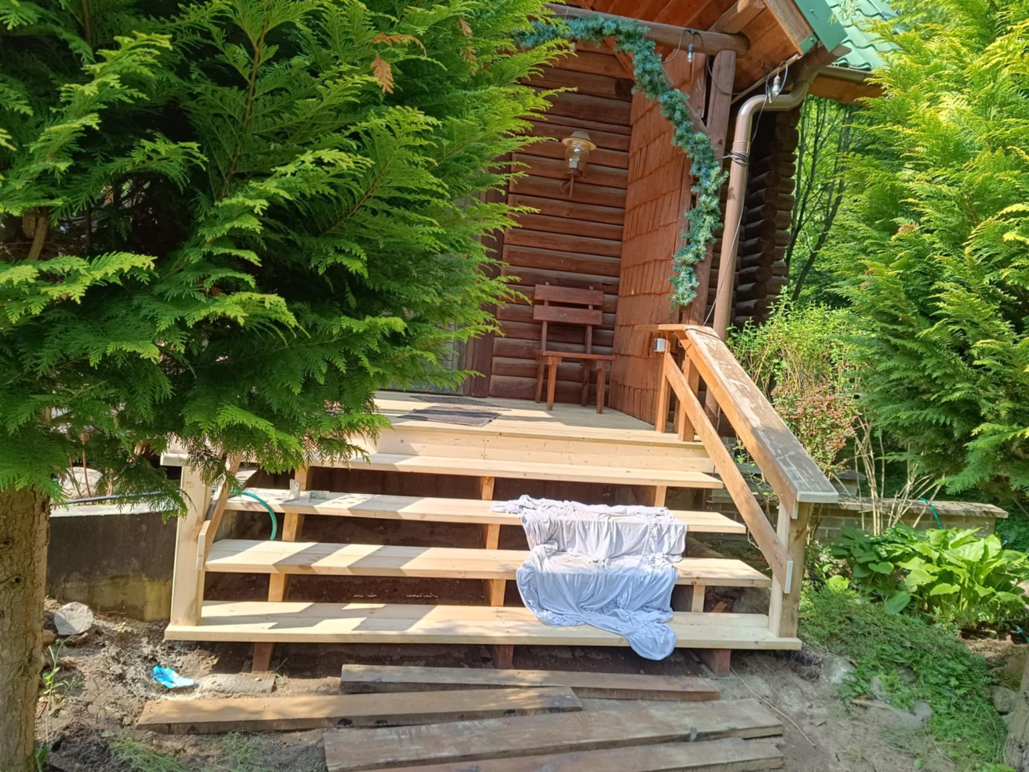 Surowe, drewniane schody prowadzące do domku z bali, otoczone zielenią. Widoczne niedokończone deski i materiał zabezpieczający, sugerujące etap budowy lub remontu.