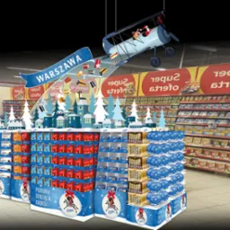 Ekspozycja promocyjna w supermarkecie z motywem zimowym i samolotem z Mikołajem, na tle regałów sklepowych; napis 'Warszawa' na banerze, opakowania produktów z motywem świątecznym.