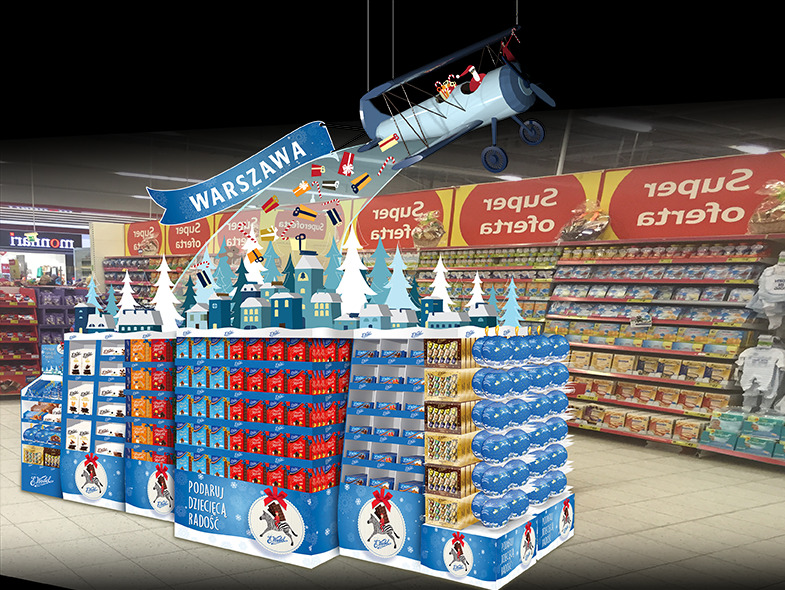 Ekspozycja promocyjna w supermarkecie z motywem zimowym i samolotem z Mikołajem, na tle regałów sklepowych; napis 'Warszawa' na banerze, opakowania produktów z motywem świątecznym.