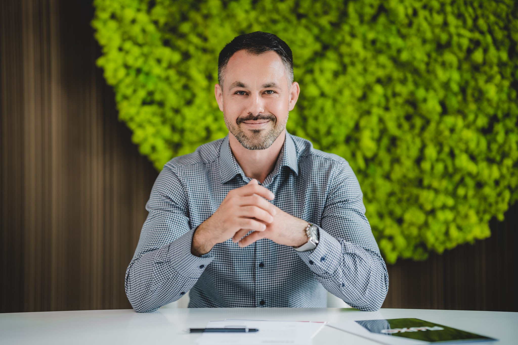 Portret mężczyzny w koszuli w drobną kratkę, siedzącego za białym biurkiem z długopisem, dokumentami i tabletem, z zieloną ścianą z mchu w tle.