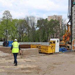 RUDBUD SP. z o.o. - Plac budowy z maszyną wiertniczą, kontenerami, przenośną toaletą i pracownikiem w żółtej kamizelce idącym w stronę robót ziemnych na tle bloku mieszkalnego.