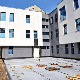 RUDBUD SP. z o.o. - Wznoszący się budynek z panelową fasadą w odcieniach bieli i szarości, widoczne okna z niebieską folią ochronną, zabezpieczona folią hydroizolacyjną płyta fundamentowa z rozłożonymi paletami...