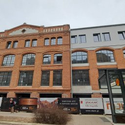 RUDBUD SP. z o.o. - Odrestaurowany budynek z czerwonej cegły z nową dobudówką, reklamy sprzedaży loftów w tle, widok z poziomu ulicy.