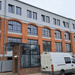 RUDBUD SP. z o.o. - Odrestaurowany budynek z czerwonej cegły z łukowymi oknami i szarą elewacją, widoczny biały samochód dostawczy i lampa uliczna.