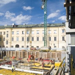 RUDBUD SP. z o.o. - Widok z góry na plac budowy z widocznymi pracami fundamentowymi, żółtymi szalunkami, zbrojeniami i pracownikami w kamizelkach odblaskowych na tle budynku i żurawia budowlanego.