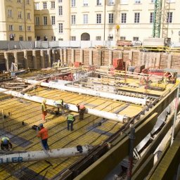 RUDBUD SP. z o.o. - Rozległy plac budowy z widocznym wykopem zabezpieczonym drewnianymi ściankami, zbrojenie fundamentów w żółtych szalunkach, pracownicy w kaskach i kamizelkach odblaskowych oraz rury do wylewania...