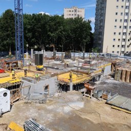 RUDBUD SP. z o.o. - Rozległy plac budowy z widocznymi fundamentami, szalunkami, zbrojeniami i pracującymi robotnikami w kaskach; w tle wysokie budynki mieszkalne i błękitne niebo z chmurami.