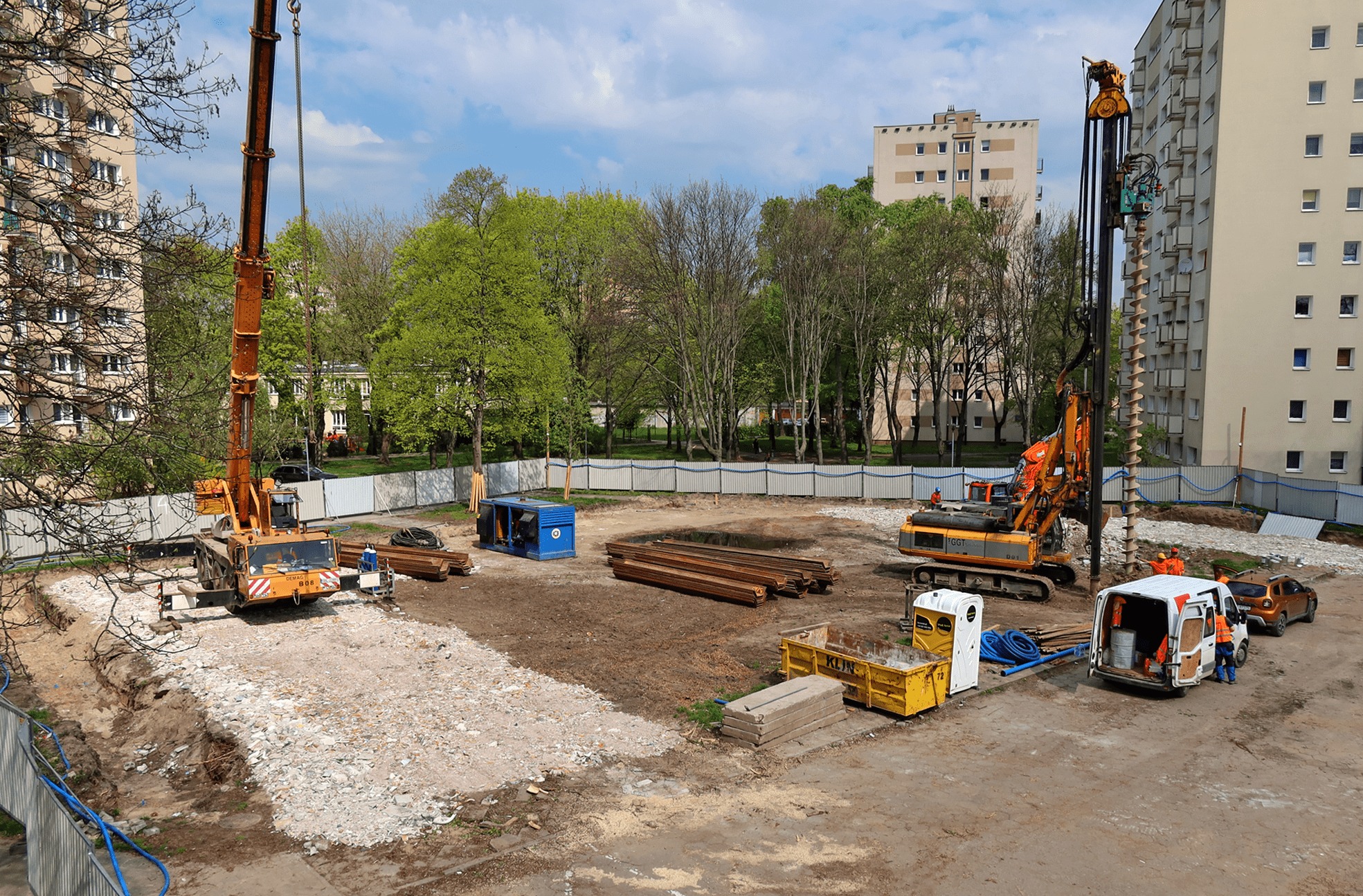 Przygotowanie terenu pod budowę: widok z góry na plac budowy z ciężkim sprzętem, w tym żurawiem i wiertnicą, oraz przygotowanym wykopem pod fundamenty, z widocznymi zbrojeniami i gruzem, na tle...