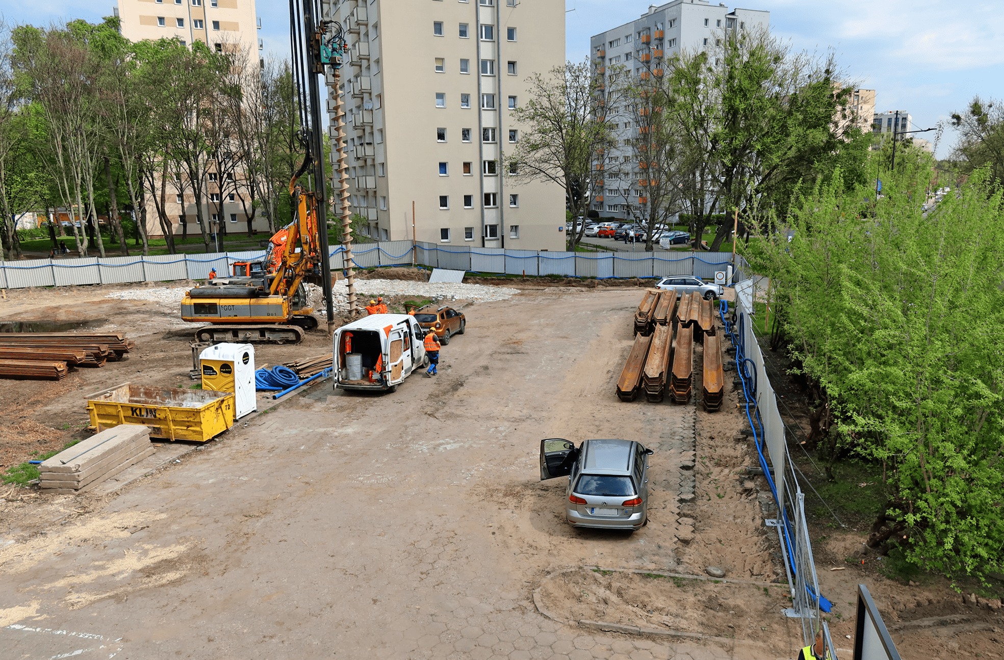 Plac budowy w miejskiej scenerii z maszyną wiertniczą, stertą stalowych kształtowników i pracownikami w pomarańczowych kamizelkach, widok z góry.
