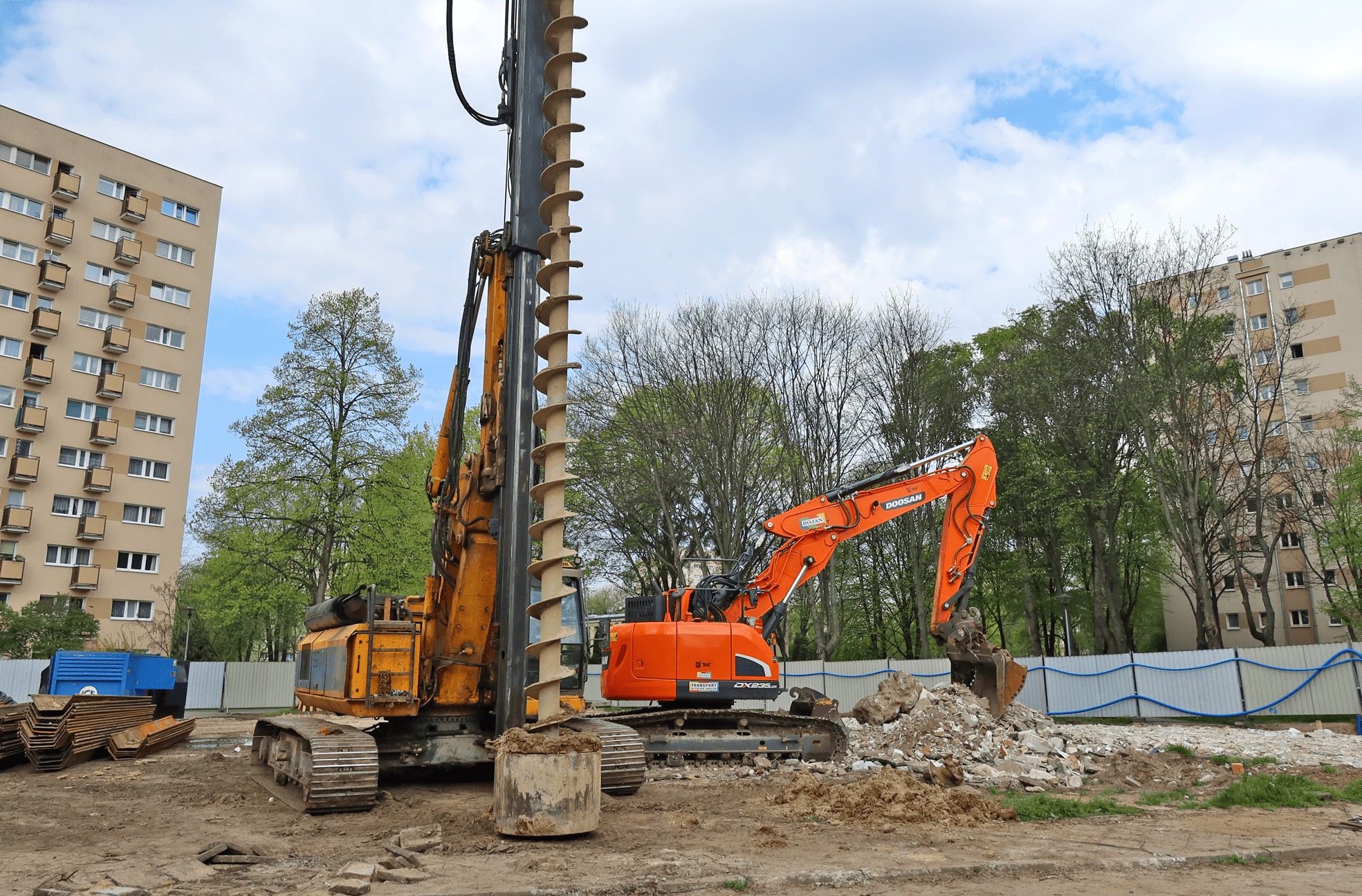 Żółta wiertnica obrotowa i pomarańczowa koparka Doosan DX235LCR na placu budowy z gruzem i ziemią, w tle budynek mieszkalny i drzewa.