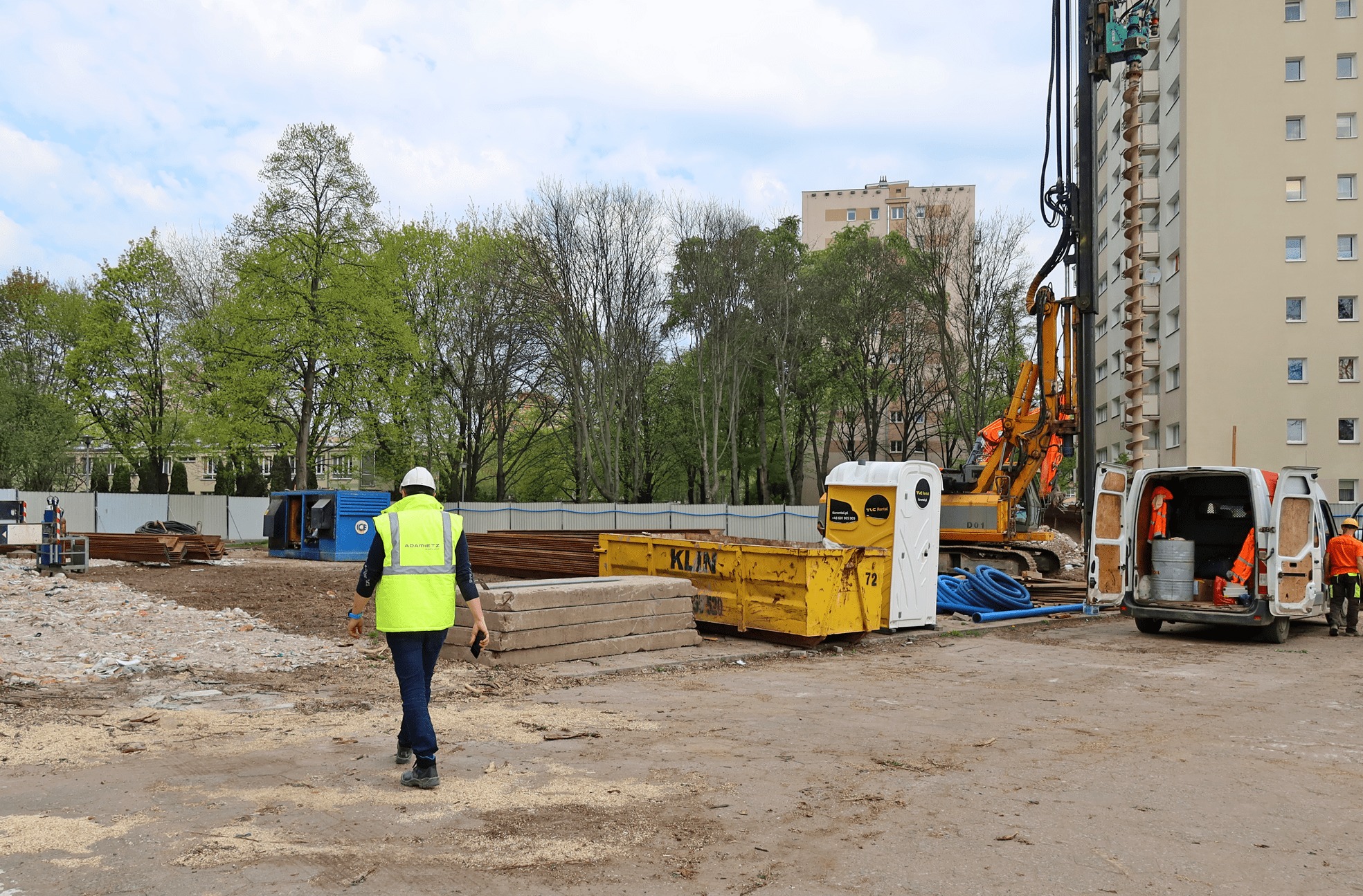 Plac budowy z maszyną wiertniczą, kontenerami, przenośną toaletą i pracownikiem w żółtej kamizelce idącym w stronę robót ziemnych na tle bloku mieszkalnego.