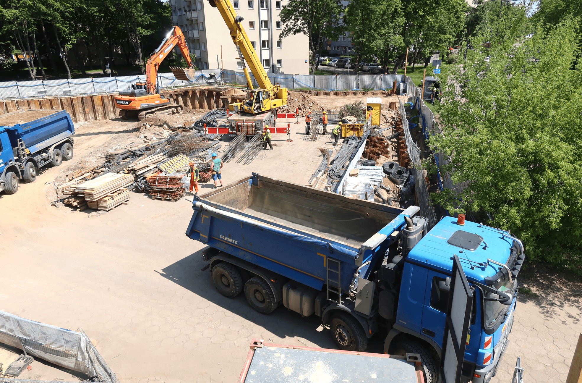 Widok z góry na plac budowy z wykopem zabezpieczonym ścianami Larsena, dźwigiem i koparką w akcji, pracownikami w kamizelkach oraz ciężarówkami Schmitz, z widocznymi zbrojeniami i deskami...