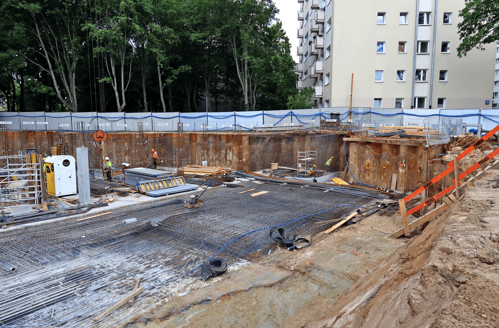 Szeroki wykop zabezpieczony ściankami Larsena, z widocznym zbrojeniem przygotowanym do zalania betonem, pracownicy w kaskach w tle, przenośna toaleta na placu budowy.