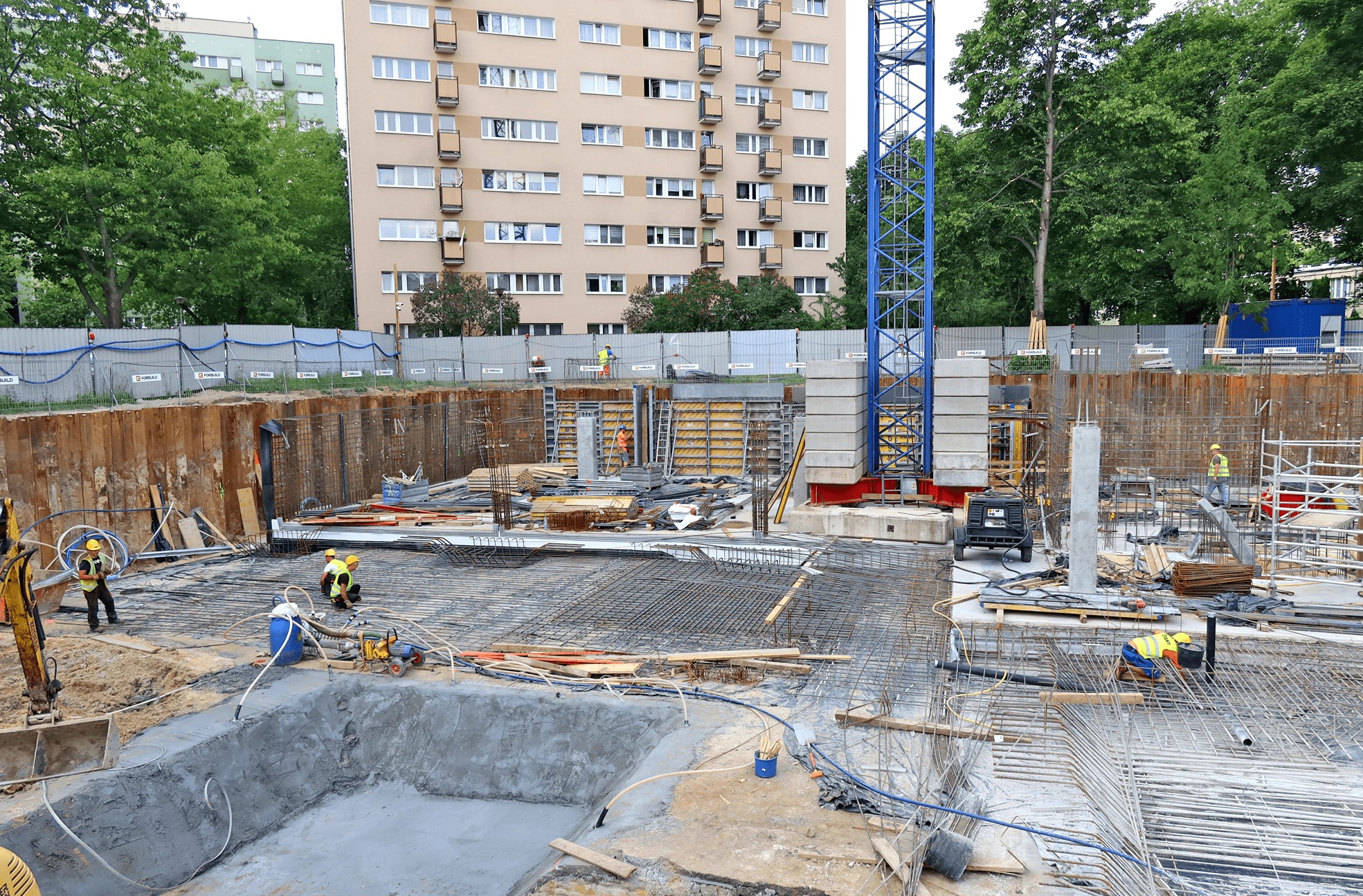 Szeroki kadr na plac budowy z wykopem pod fundamenty, zbrojeniami, betonową wylewką w kształcie basenu, pracownikami w żółtych kamizelkach i niebieskim żurawiem w tle oraz budynkiem mieszkalnym...