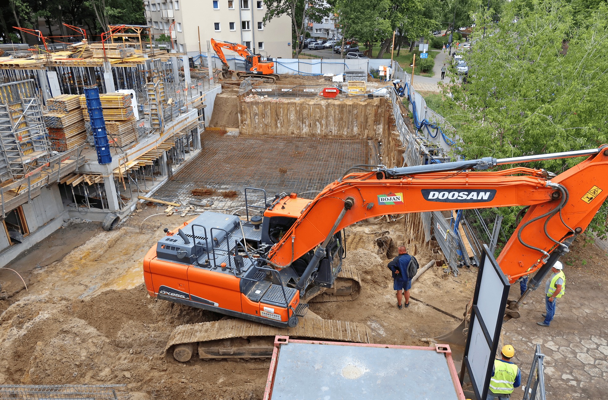 Widok z góry na plac budowy z dwoma pomarańczowymi koparkami Doosan, wykopem zabezpieczonym ściankami Larsena, zbrojeniem fundamentów i częściowo wzniesioną konstrukcją budynku.
