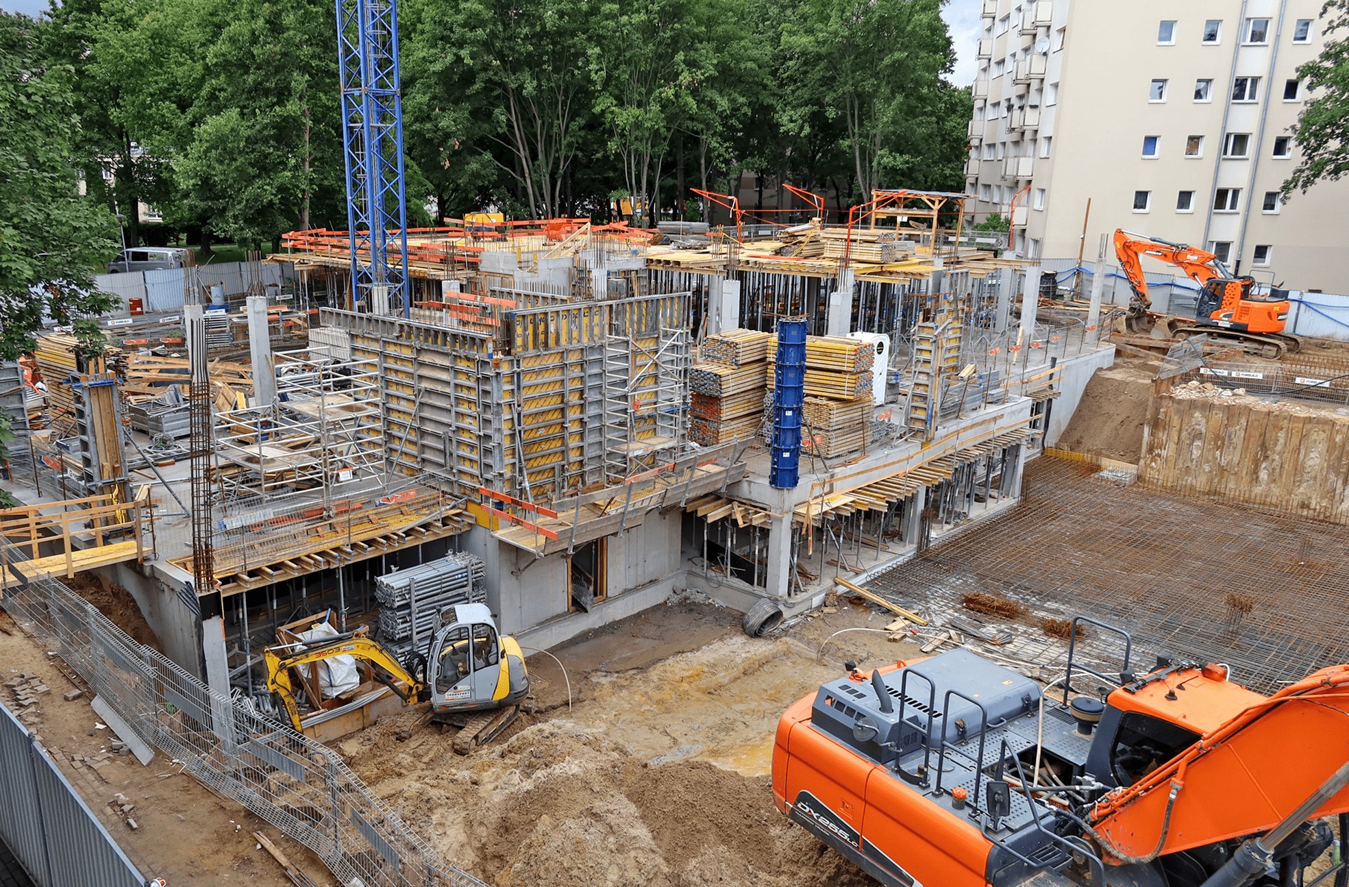 Rozległy plac budowy z widocznymi szalunkami, zbrojeniami, wykopami i ciężkim sprzętem, w tym pomarańczową koparką Hitachi DX265LC na pierwszym planie i żółtą mini-koparką w tle, z budynkami...