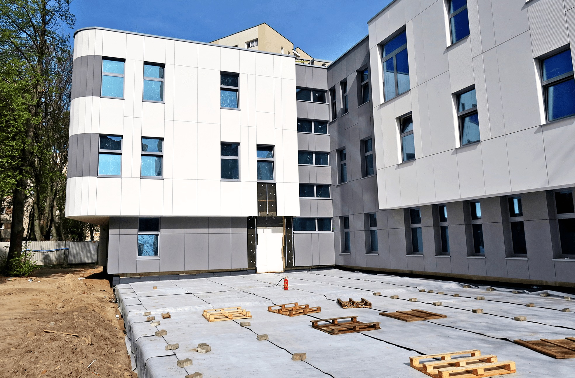 Wznoszący się budynek z panelową fasadą w odcieniach bieli i szarości, widoczne okna z niebieską folią ochronną, zabezpieczona folią hydroizolacyjną płyta fundamentowa z rozłożonymi paletami...