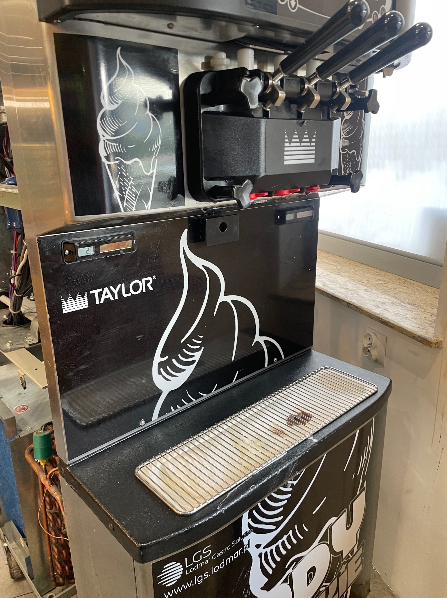 Profesjonalna maszyna do lodów Taylor z trzema dyszami, ozdobiona grafiką lodów i logo firmy, z widocznym panelem sterowania i tacką ociekową.