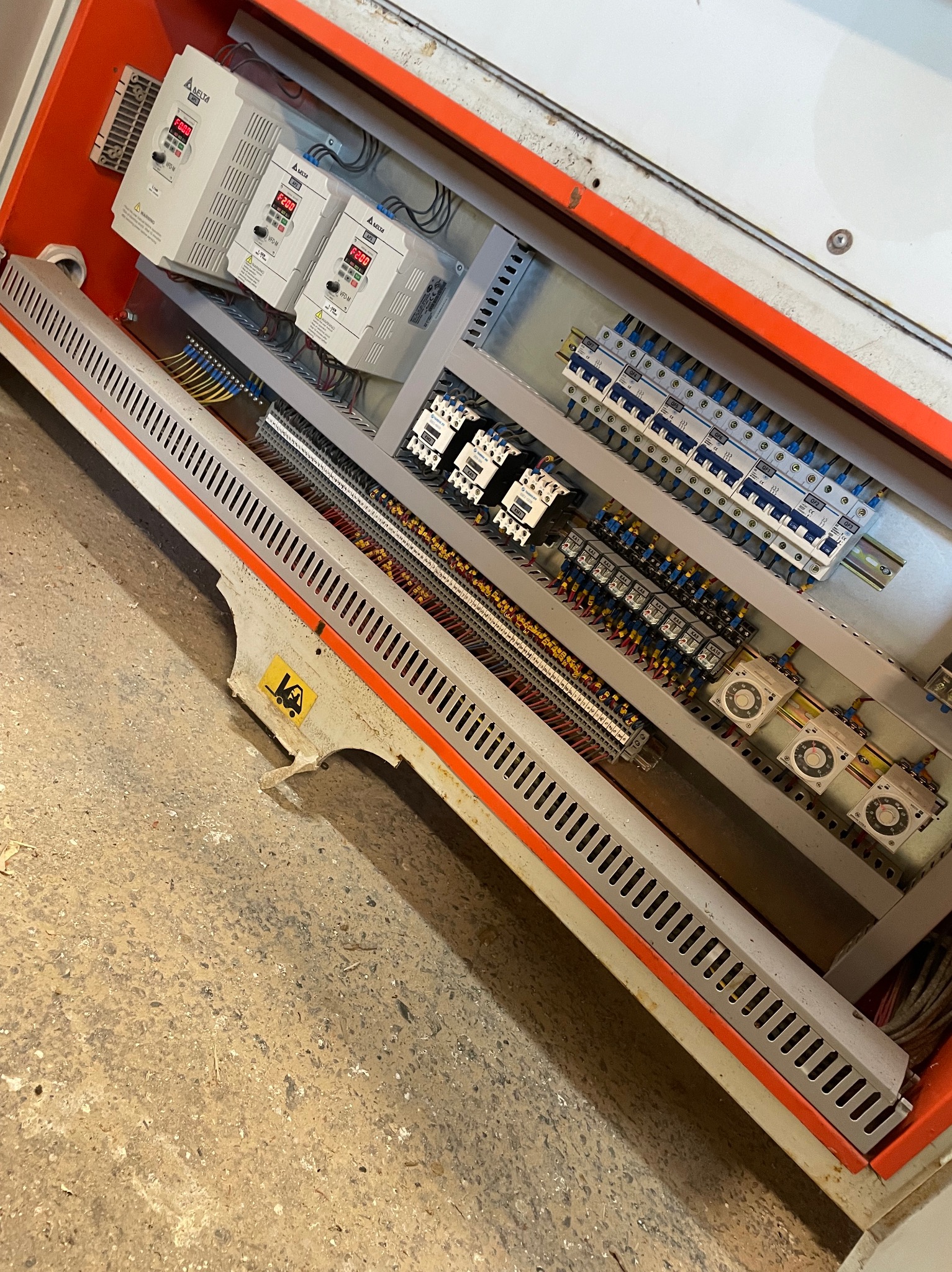 Otwarta szafa sterownicza maszyny z widocznymi elementami elektrycznymi, w tym falownikami Delta VFD-M, bezpiecznikami, przekaźnikami i okablowaniem, umieszczona w pomarańczowej obudowie z metalową...