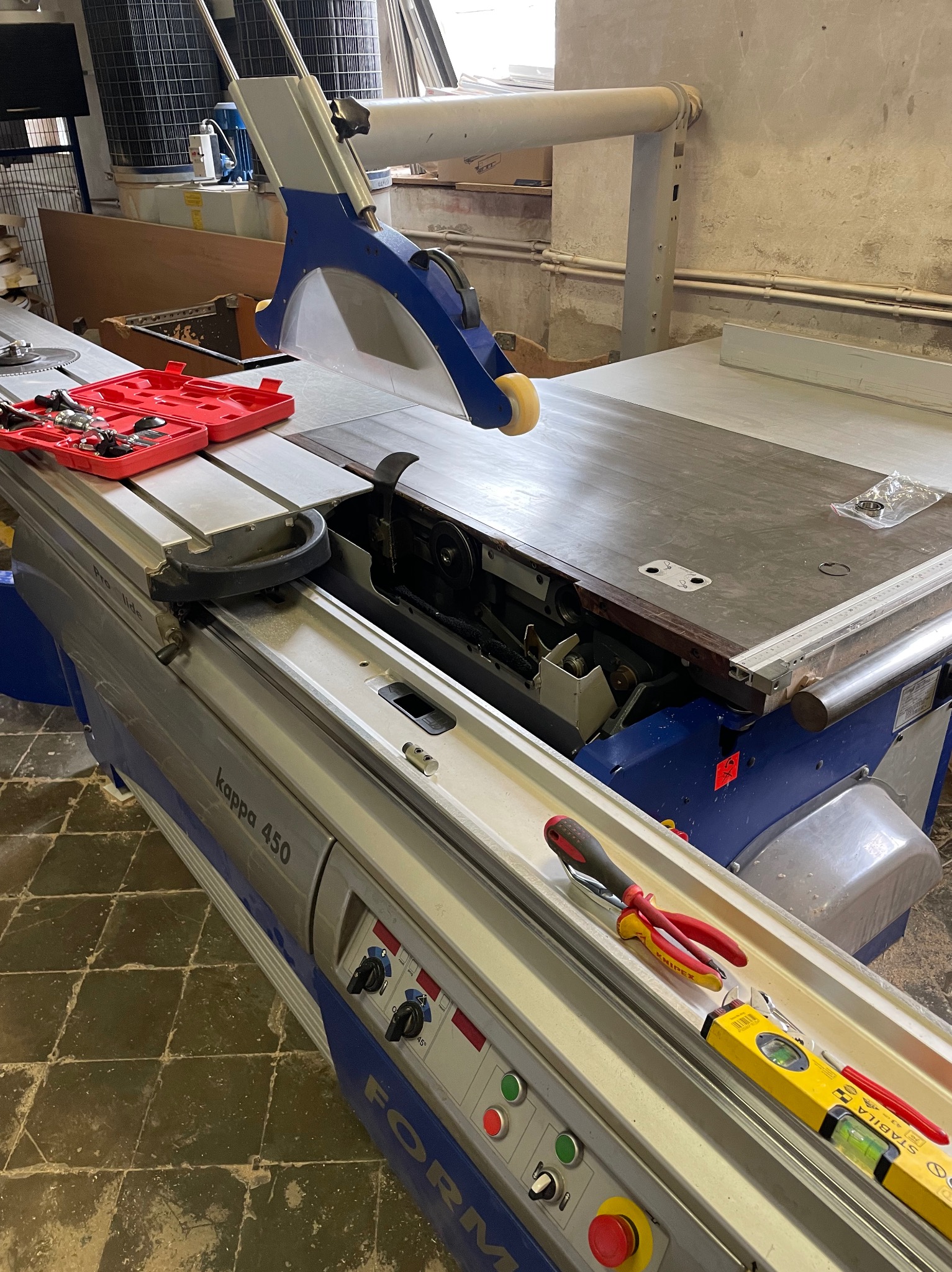 Piła formatowa Kappa 450 z otwartą obudową tarczy, obok zestaw narzędzi w czerwonym pudełku i poziomica Stabila, na blacie leżą części zamienne w folii.