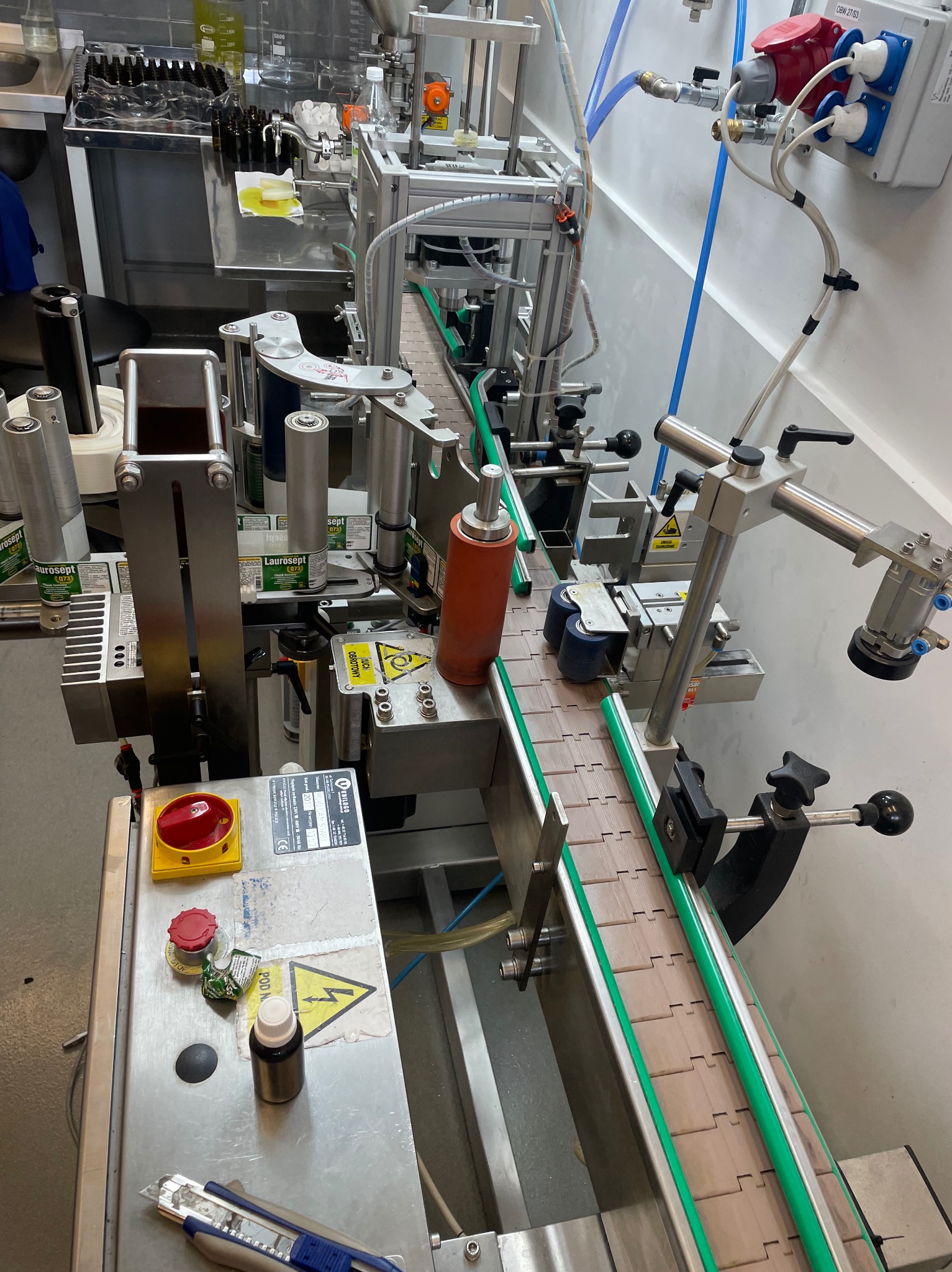 Automatyczna linia produkcyjna z systemem transportującym butelki, na której widoczne są elementy etykietujące i dozujące, z bliska widoczny panel sterowania z przyciskiem awaryjnym i butelką...