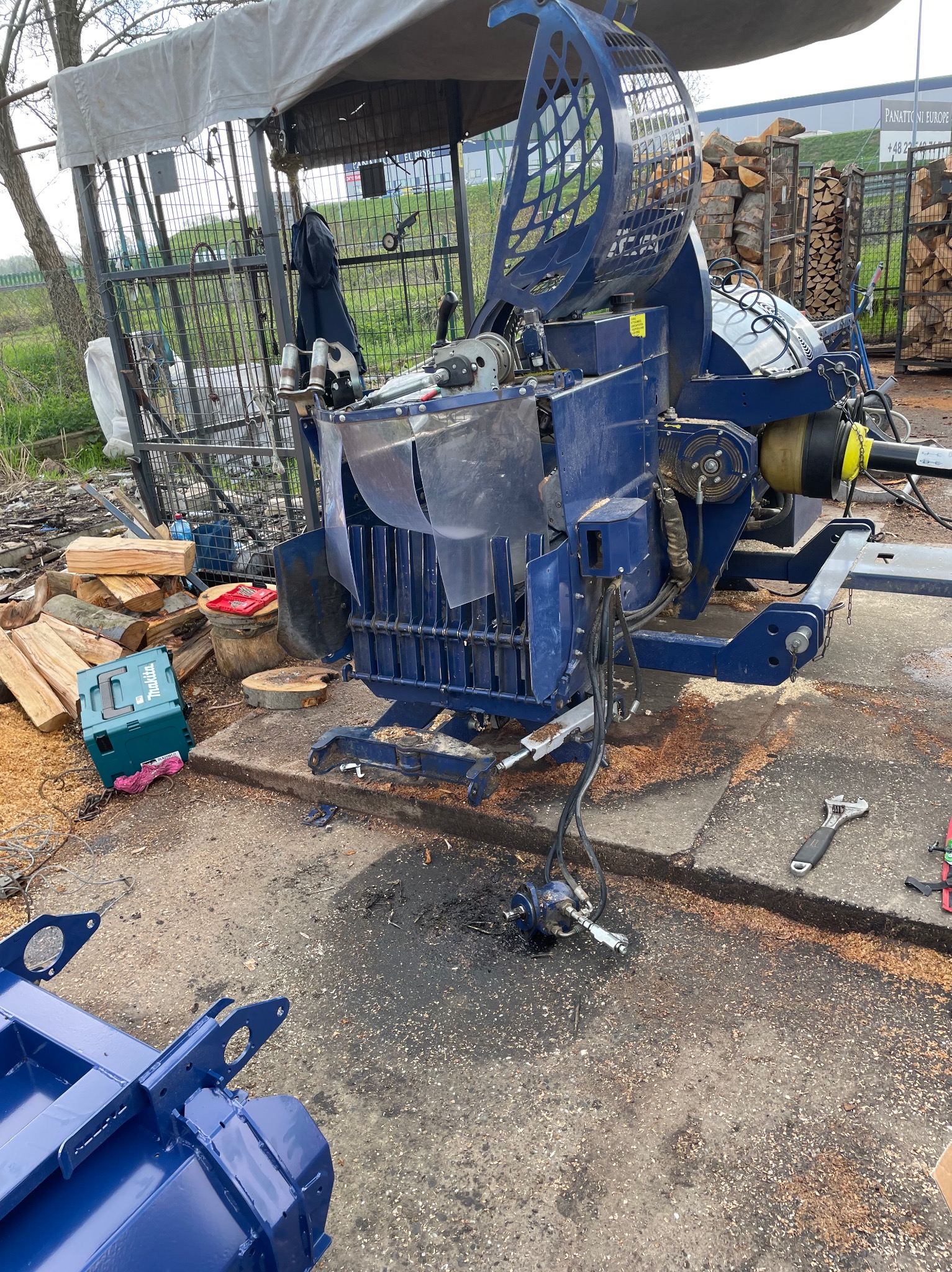 Niebieska, hydrauliczna łuparka do drewna z osłoną, skrzynką narzędziową Makita i stosem pociętych klocków drewna w tle, na zewnątrz.