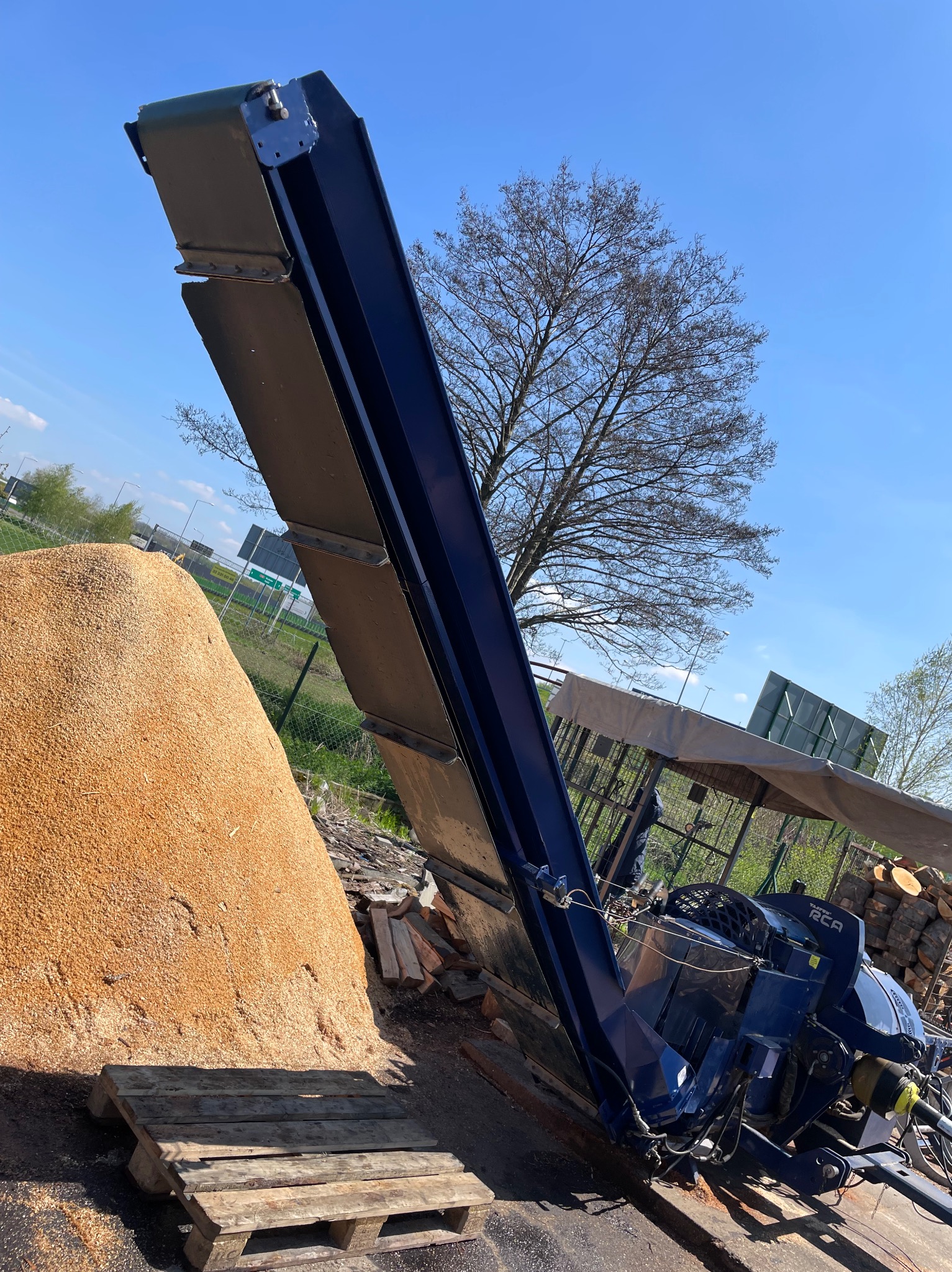 Niebieska maszyna do obróbki drewna z podajnikiem taśmowym, obok pryzma trocin na palecie, w tle drzewo bez liści i błękitne niebo.