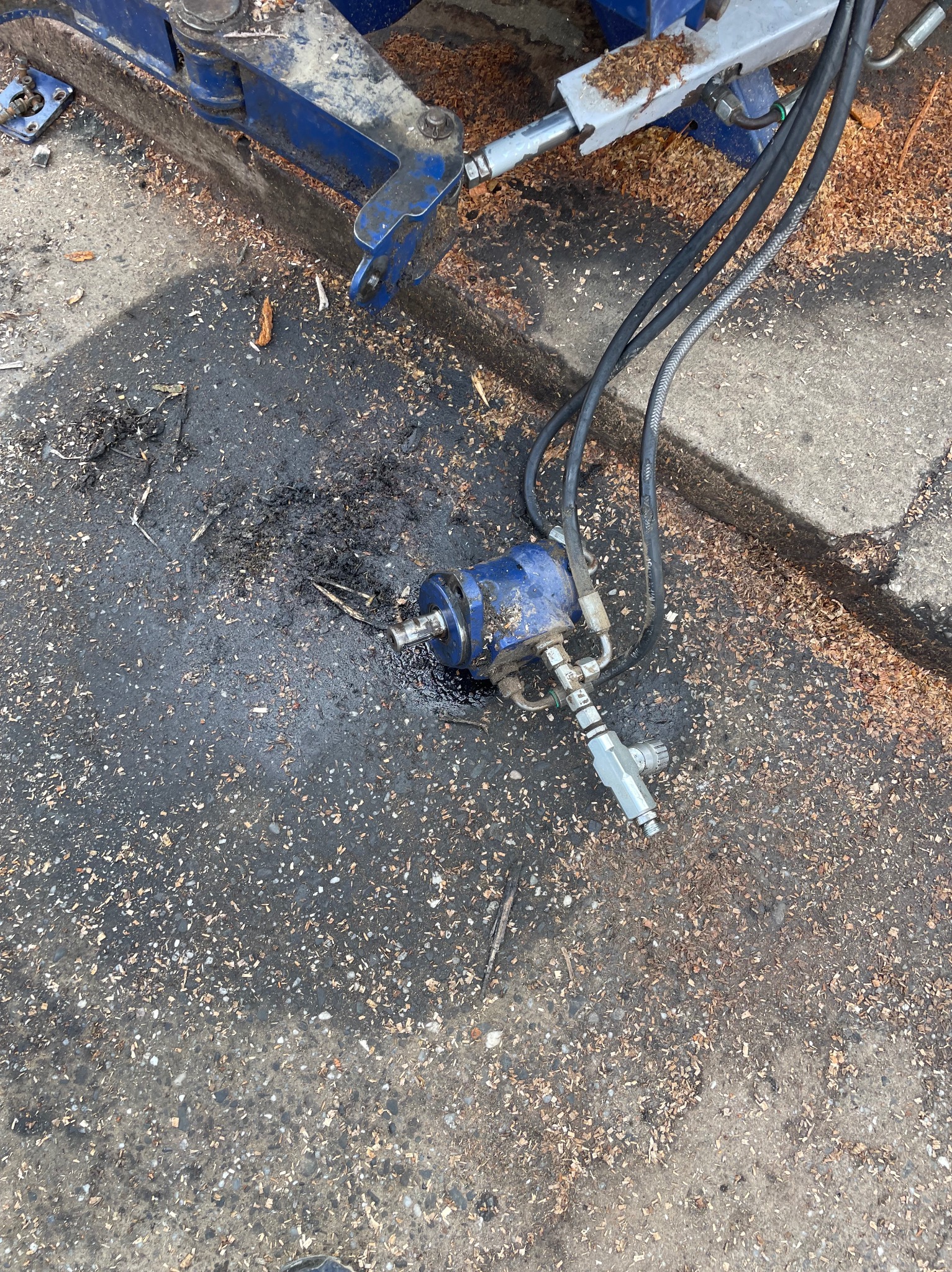 Wyciek oleju z niebieskiego elementu hydraulicznego maszyny na brudnym betonowym podłożu pokrytym trocinami, widoczne przewody hydrauliczne i fragment ramy maszyny.