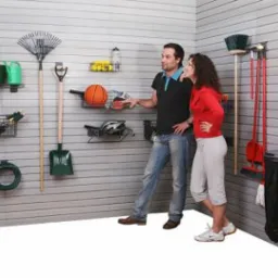 Zabudowa garażu z systemem paneli ściennych z haczykami i półkami, na których zawieszone są narzędzia ogrodnicze, sprzęt sportowy i akcesoria, prezentowana przez parę stojącą w rogu pomieszczenia.