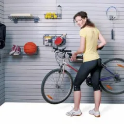Kobieta umieszcza rower na ścianie z panelami, na której wiszą kosze z piłką, butami, rakietą do badmintona i innymi przedmiotami, zabudowa garażu.