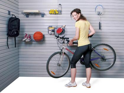 Kobieta umieszcza rower na ścianie z panelami, na której wiszą kosze z piłką, butami, rakietą do badmintona i innymi przedmiotami, zabudowa garażu.