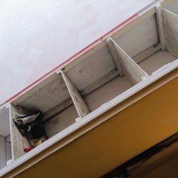 Klimek maluje - Widok z dołu na drewniany regał z półkami, świeżo pomalowany na biało, z widocznymi śladami farby na ścianie w tle, w jednej z przegródek leżą robocze rękawice.