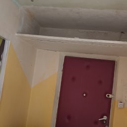 Klimek maluje - Pokój w trakcie odświeżania: sufit z widocznymi śladami gładzi, wisząca żarówka na kablu, częściowo pomalowane ściany w kolorach żółtym i białym, drzwi wejściowe w odcieniu bordo.